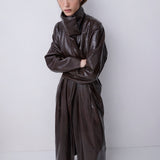 A model wearing a dark brown, high-collared, wide-leg faux-leather coat with a textured finish, standing against a plain light gray studio background.