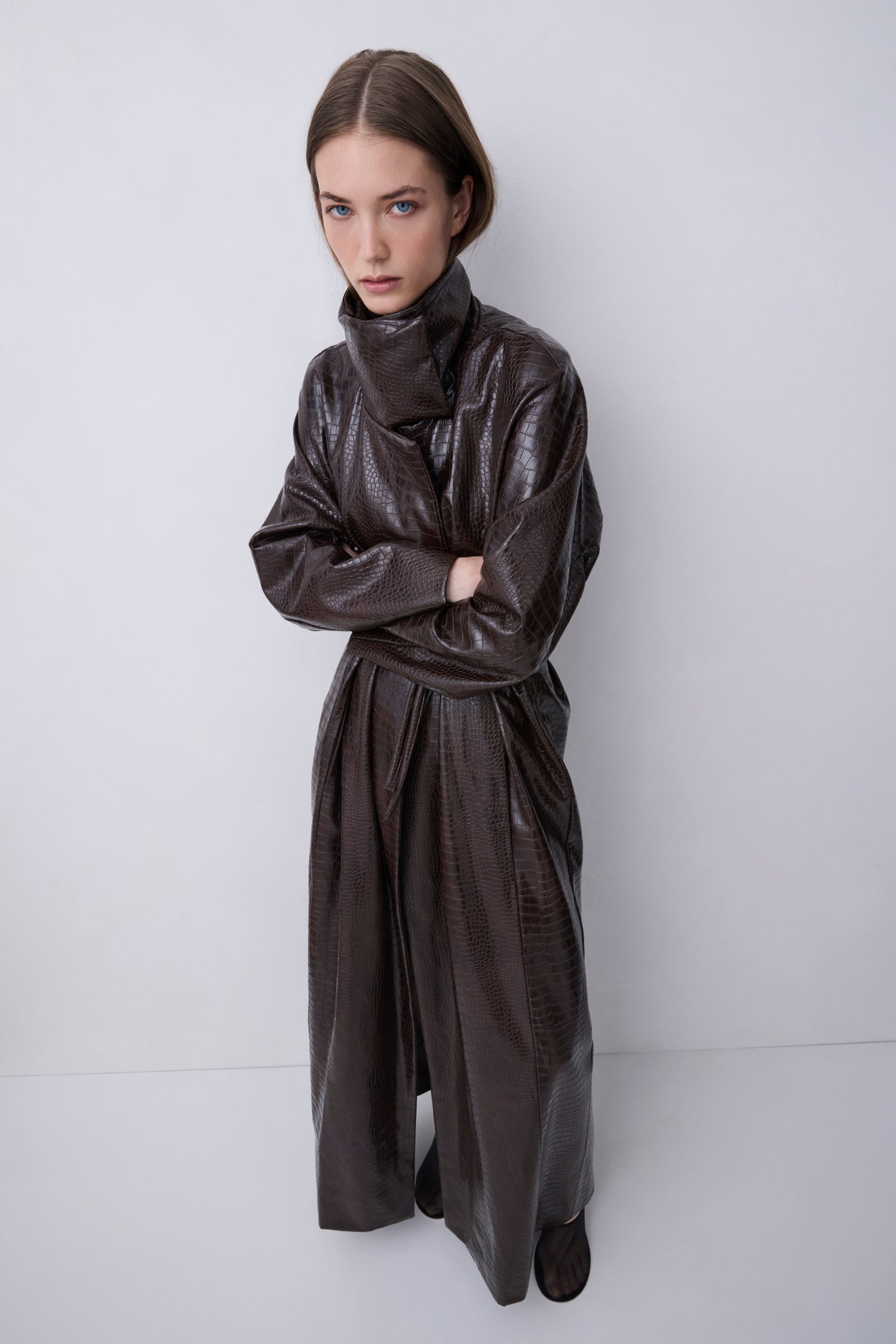 A model wearing a dark brown, high-collared, wide-leg faux-leather coat with a textured finish, standing against a plain light gray studio background.