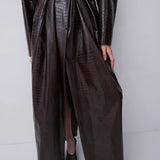 A model wearing a dark brown faux-crocodile textured trench coat with a matching tie belt, paired with black pointed-toe shoes, against a plain light gray studio background.