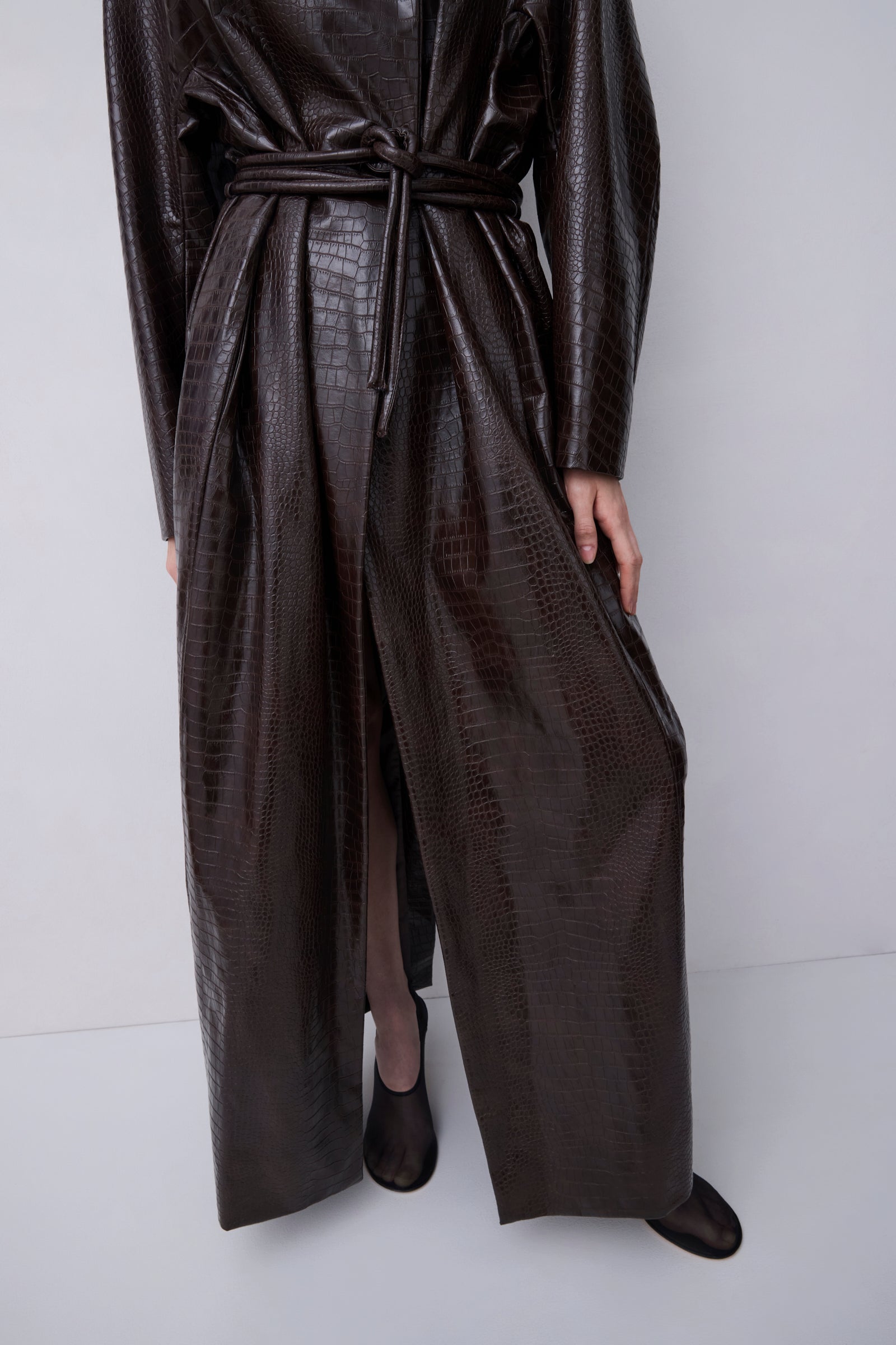 A model wearing a dark brown faux-crocodile textured trench coat with a matching tie belt, paired with black pointed-toe shoes, against a plain light gray studio background.