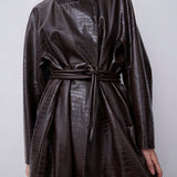 A model wearing a dark brown faux-crocodile leather high-neck wrap coat with a matching tie belt, shown from the neck down against a neutral light gray background.