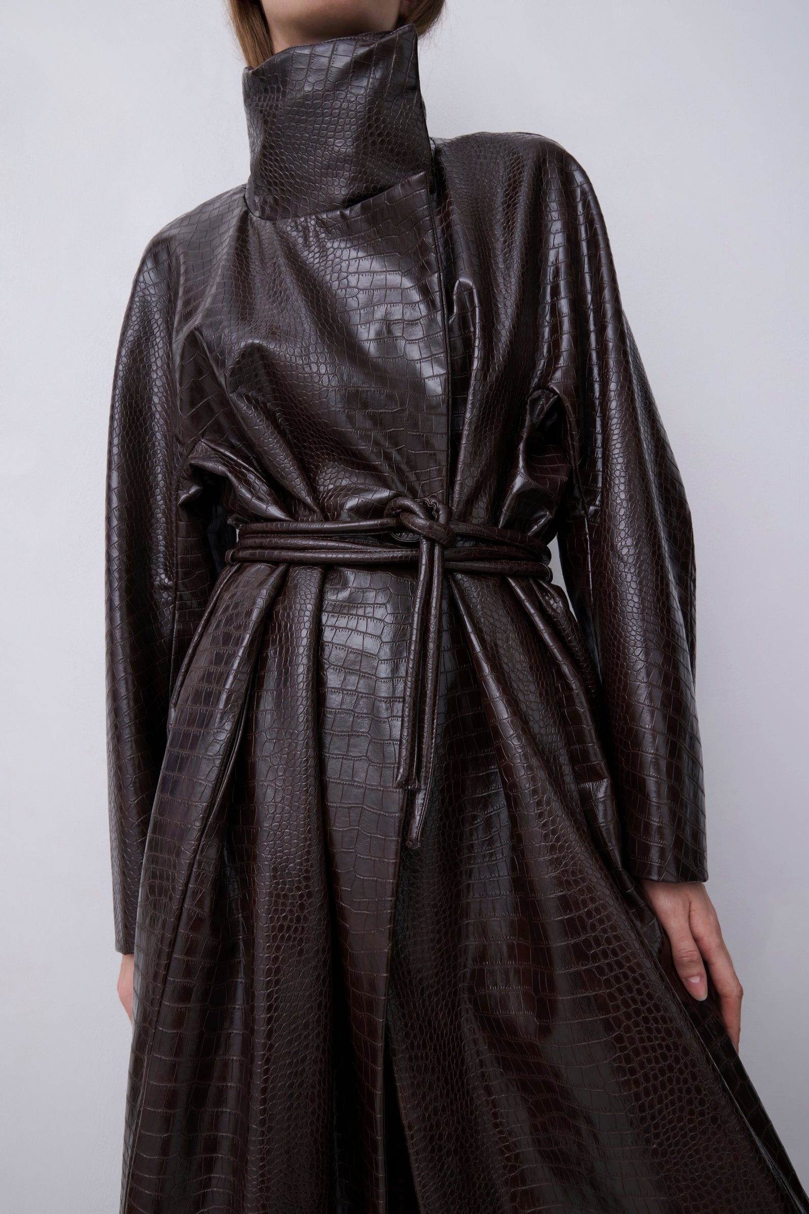 A model wearing a dark brown faux-crocodile leather high-neck wrap coat with a matching tie belt, shown from the neck down against a neutral light gray background.