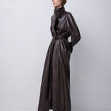 A model wearing a dark brown, glossy, textured faux-croc long coat with a high collar and waist tie, standing against a light gray studio background.