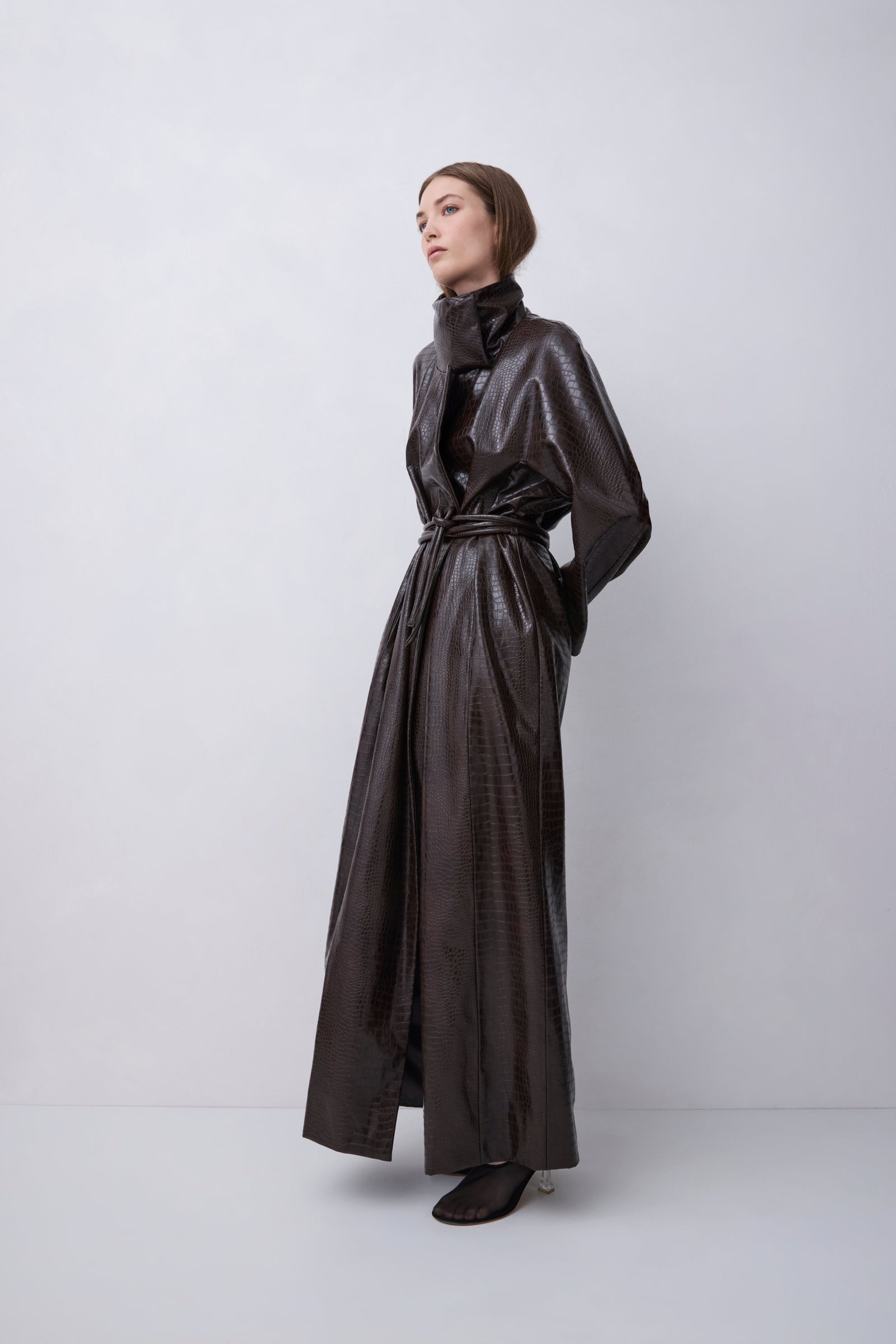 A model wearing a dark brown, glossy, textured faux-croc long coat with a high collar and waist tie, standing against a light gray studio background.