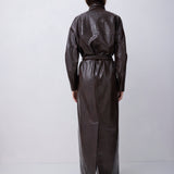 A model viewed from the back wearing a dark brown, glossy, textured long coat or dress with a tie belt, standing against a plain light gray studio background.