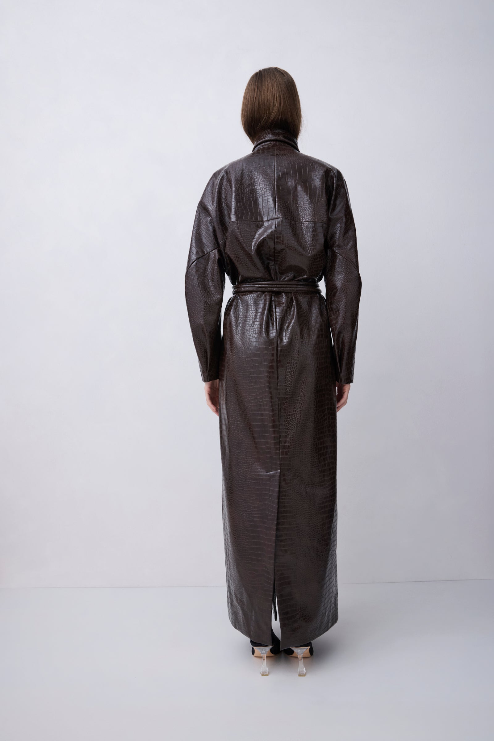 A model viewed from the back wearing a dark brown, glossy, textured long coat or dress with a tie belt, standing against a plain light gray studio background.