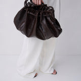 A person wearing white wide-leg pants holds a dark brown textured leather drawstring bag against a clean white studio background.