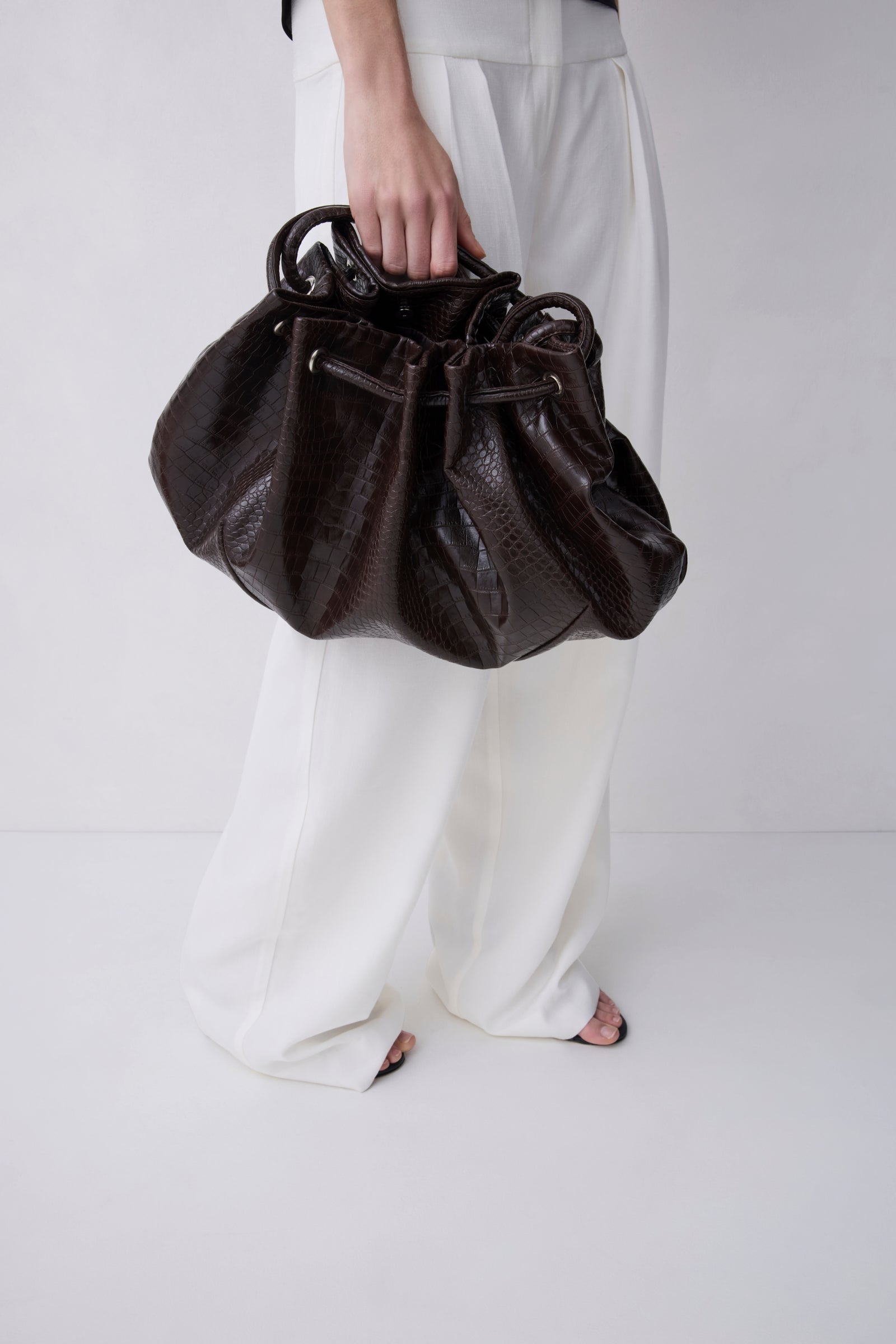 A person wearing white wide-leg pants holds a dark brown textured leather drawstring bag against a clean white studio background.
