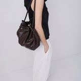 A model wearing a black sleeveless top and white wide-leg trousers carries a dark brown textured leather bucket bag over her shoulder, photographed against a clean white studio background.