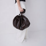 A person wearing a black top and white wide-leg pants holds a dark brown textured leather drawstring bucket bag against a plain light gray studio background.