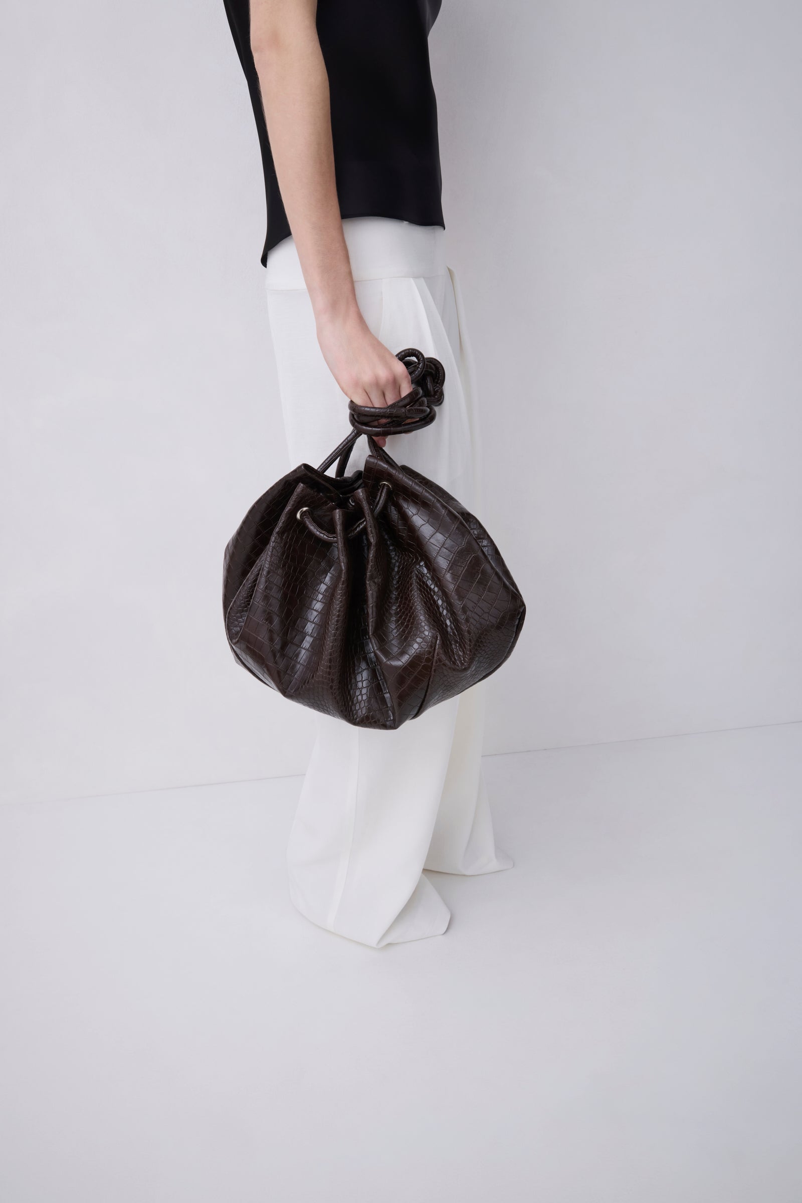 A person wearing a black top and white wide-leg pants holds a dark brown textured leather drawstring bucket bag against a plain light gray studio background.