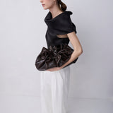 A model in a studio setting, wearing a black asymmetrical top and white wide-leg trousers, holding a dark brown gathered leather clutch bag.
