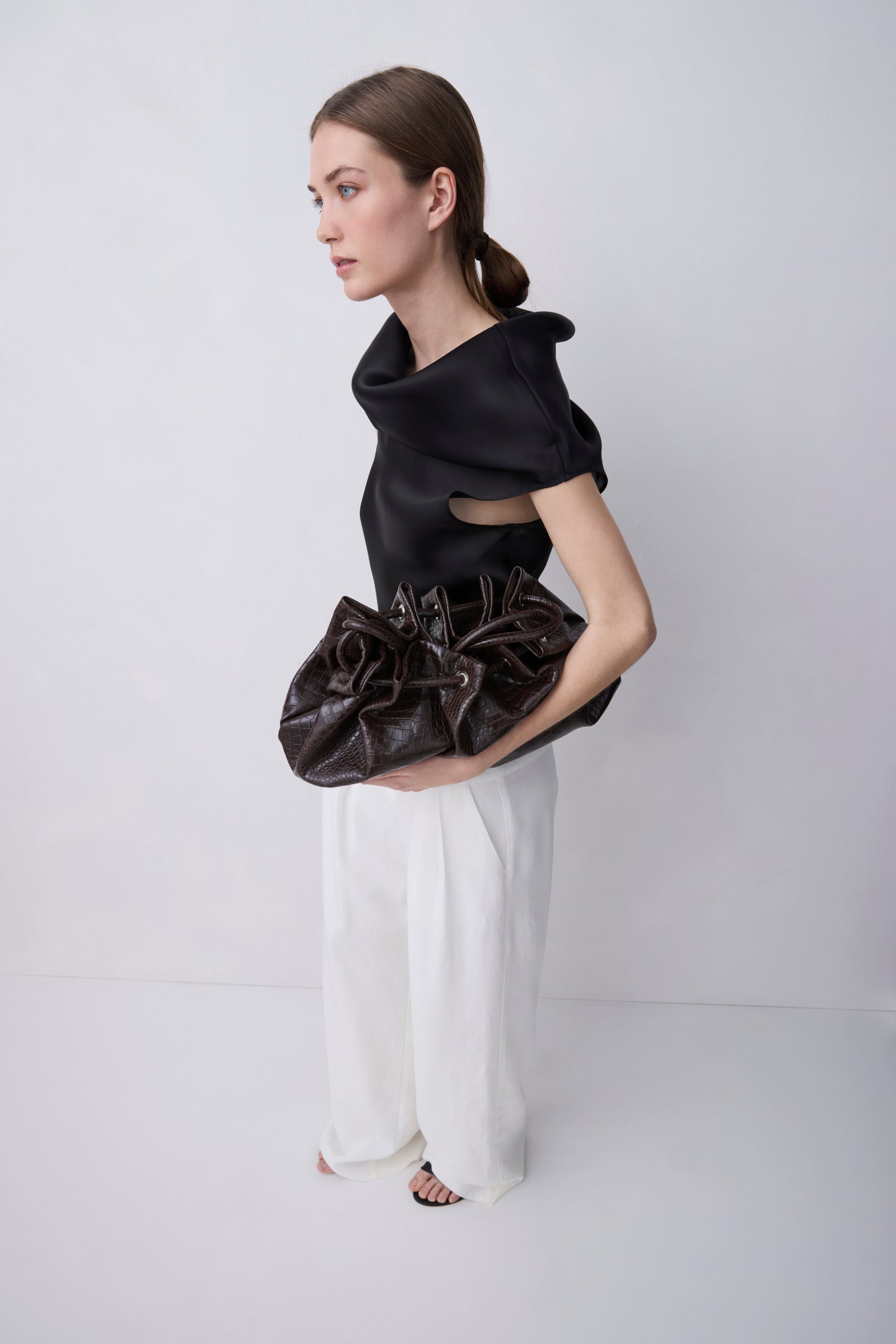 A model in a studio setting, wearing a black asymmetrical top and white wide-leg trousers, holding a dark brown gathered leather clutch bag.