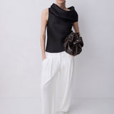 A model stands against a light gray studio background wearing a black sleeveless draped top, white wide-leg pants, and holding a dark brown crumpled leather bag.