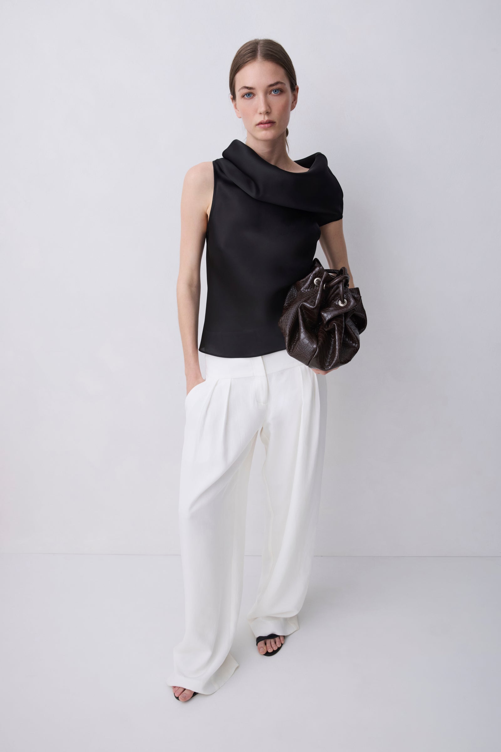 A model stands against a light gray studio background wearing a black sleeveless draped top, white wide-leg pants, and holding a dark brown crumpled leather bag.