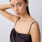A woman with dark hair pulled back wears a dark brown satin spaghetti-strap top against a light gray studio background.