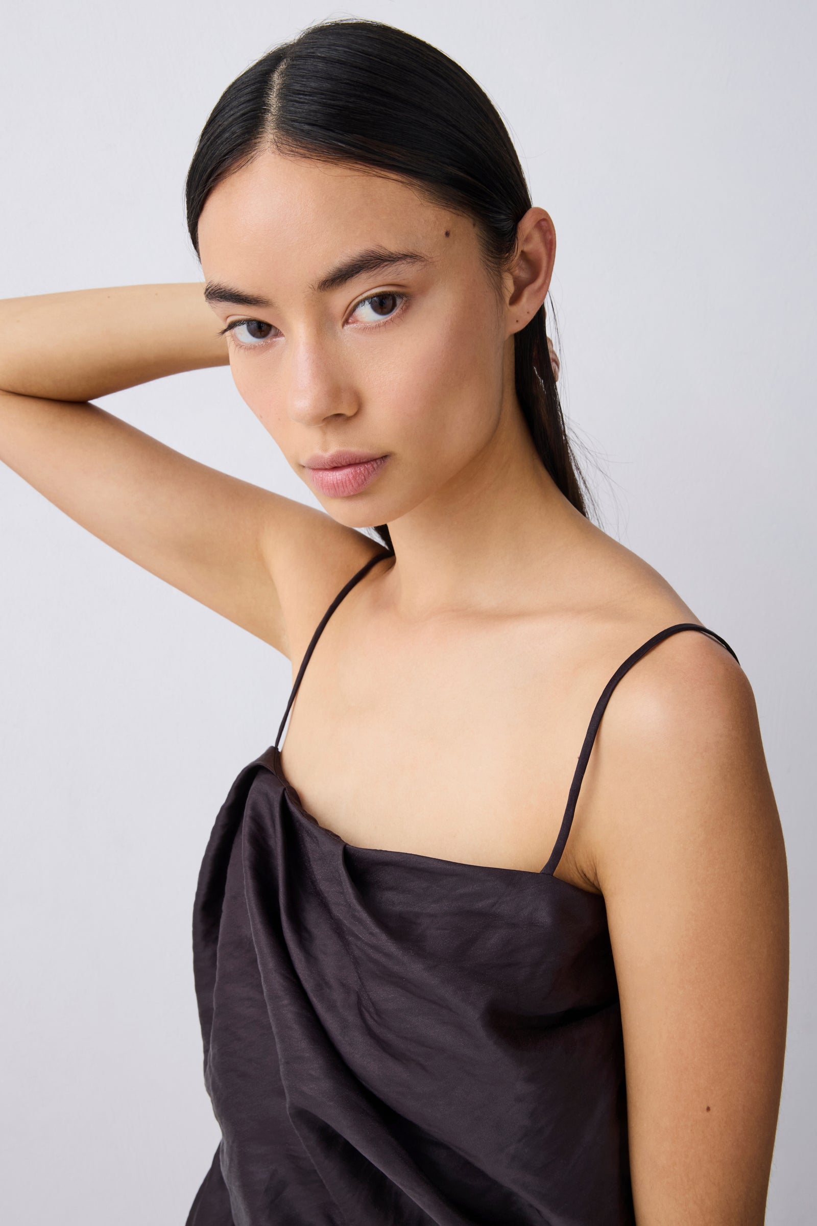 A woman with dark hair pulled back wears a dark brown satin spaghetti-strap top against a light gray studio background.