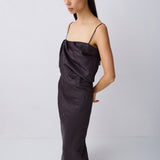 A woman modeling a dark charcoal gray satin slip dress with thin spaghetti straps and a draped, gathered bodice, paired with black flat sandals against a light gray studio background.