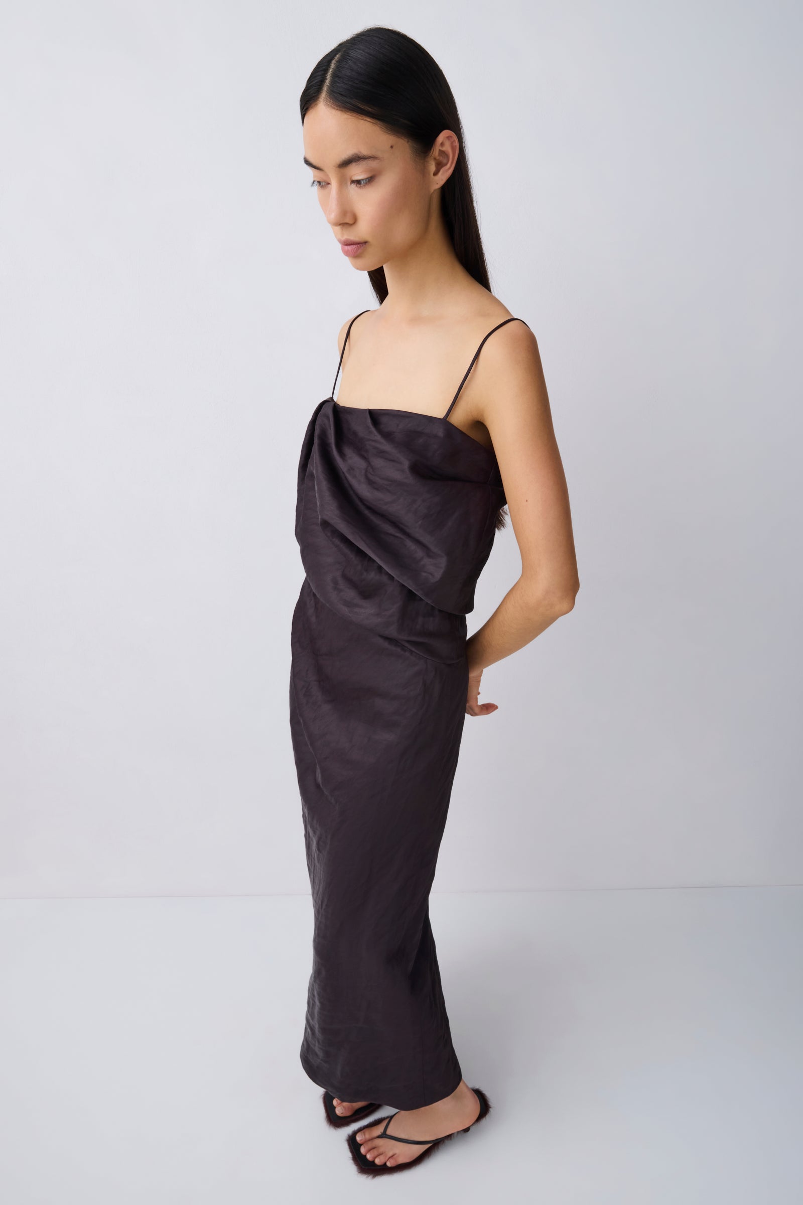 A woman modeling a dark charcoal gray satin slip dress with thin spaghetti straps and a draped, gathered bodice, paired with black flat sandals against a light gray studio background.