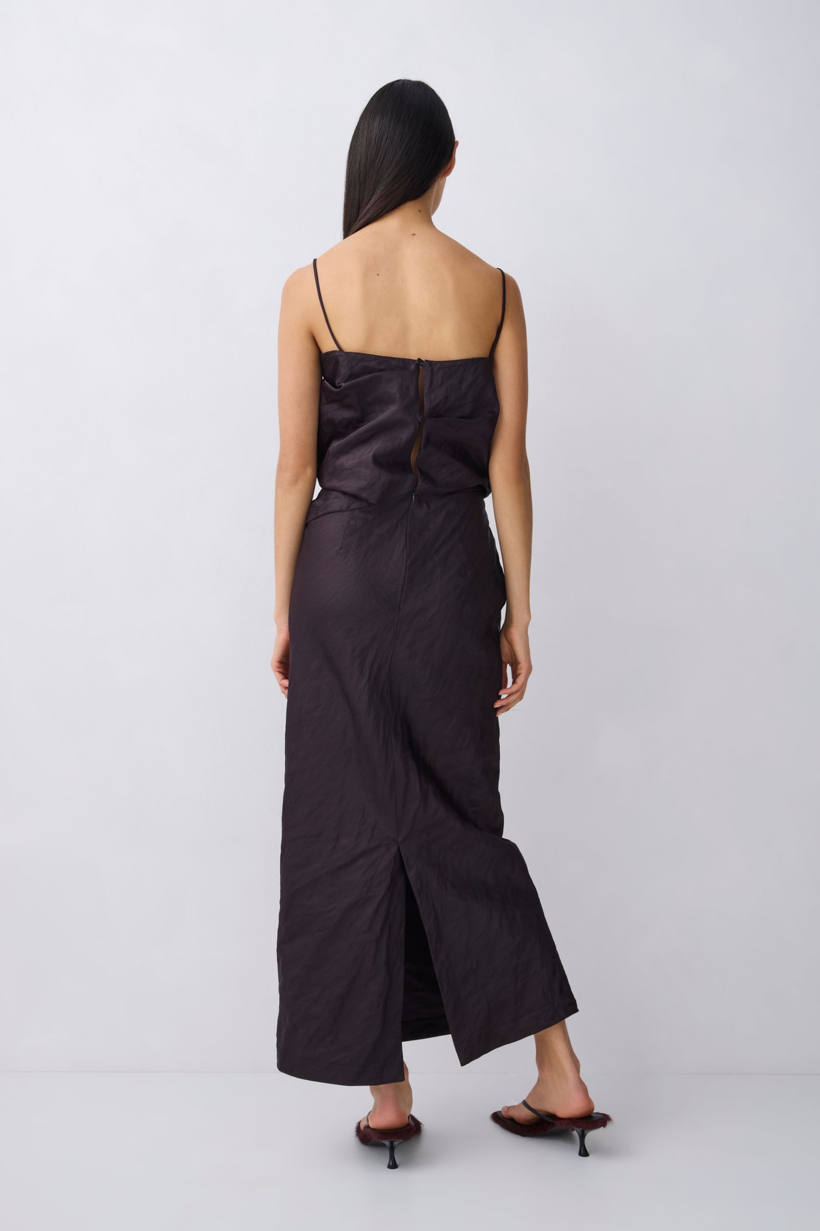 A woman viewed from the back wearing a black spaghetti-strap maxi dress with a gathered bodice and wide-leg skirt, paired with black heeled sandals, against a light gray studio background.