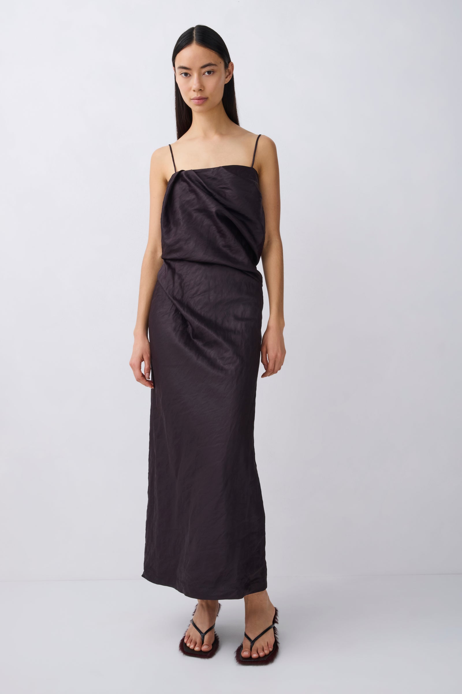 A model wearing a black draped spaghetti-strap maxi dress with a gathered waist, paired with black thong sandals, against a light gray studio background.