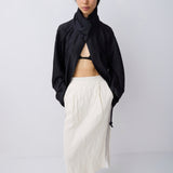 A model stands against a light gray studio background wearing a black asymmetrical draped jacket, a cream-colored pleated midi skirt, and black pointed-toe flats.