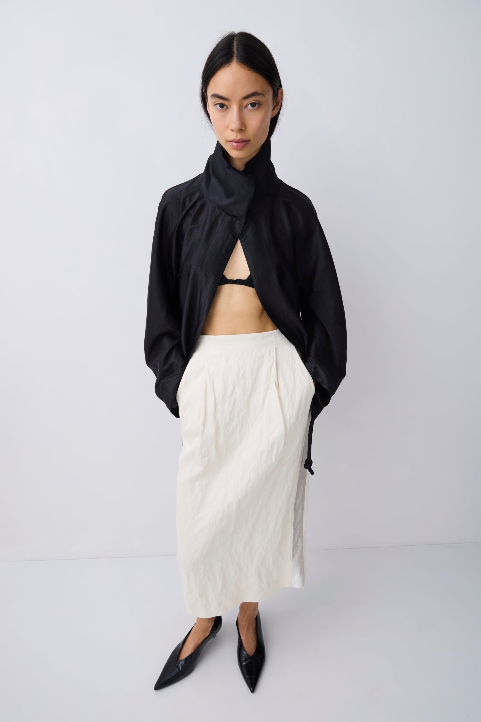 A model stands against a light gray studio background wearing a black asymmetrical draped jacket, a cream-colored pleated midi skirt, and black pointed-toe flats.