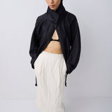 A model stands against a light gray studio background wearing a black asymmetrical high-neck jacket, an off-white textured midi skirt, and black pointed-toe heels.