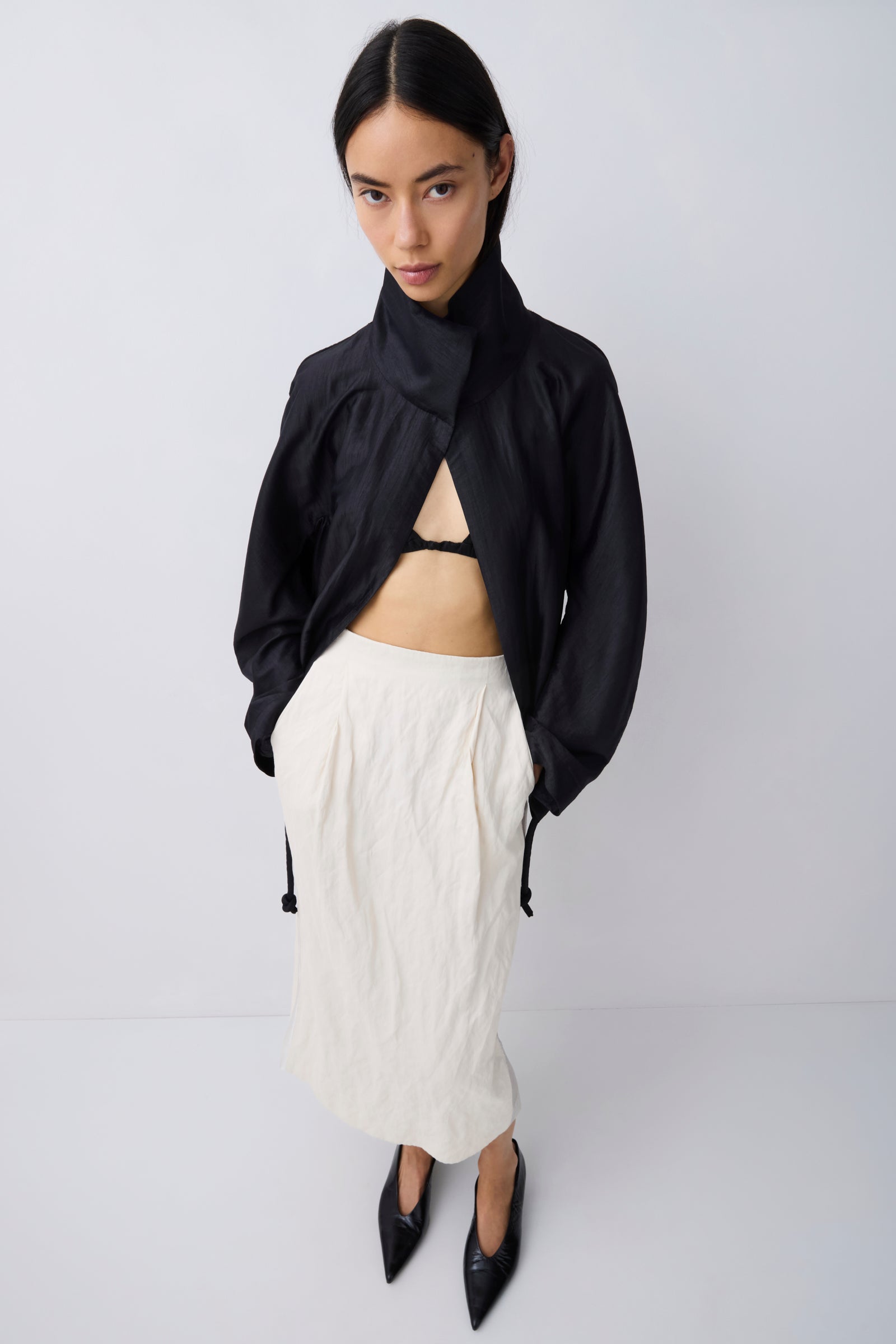 A model stands against a light gray studio background wearing a black asymmetrical high-neck jacket, an off-white textured midi skirt, and black pointed-toe heels.