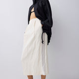 A model wearing a black cropped long-sleeve jacket with a high neck, paired with a cream-colored textured midi skirt and black pointed-toe kitten heels, set against a plain light gray studio background.