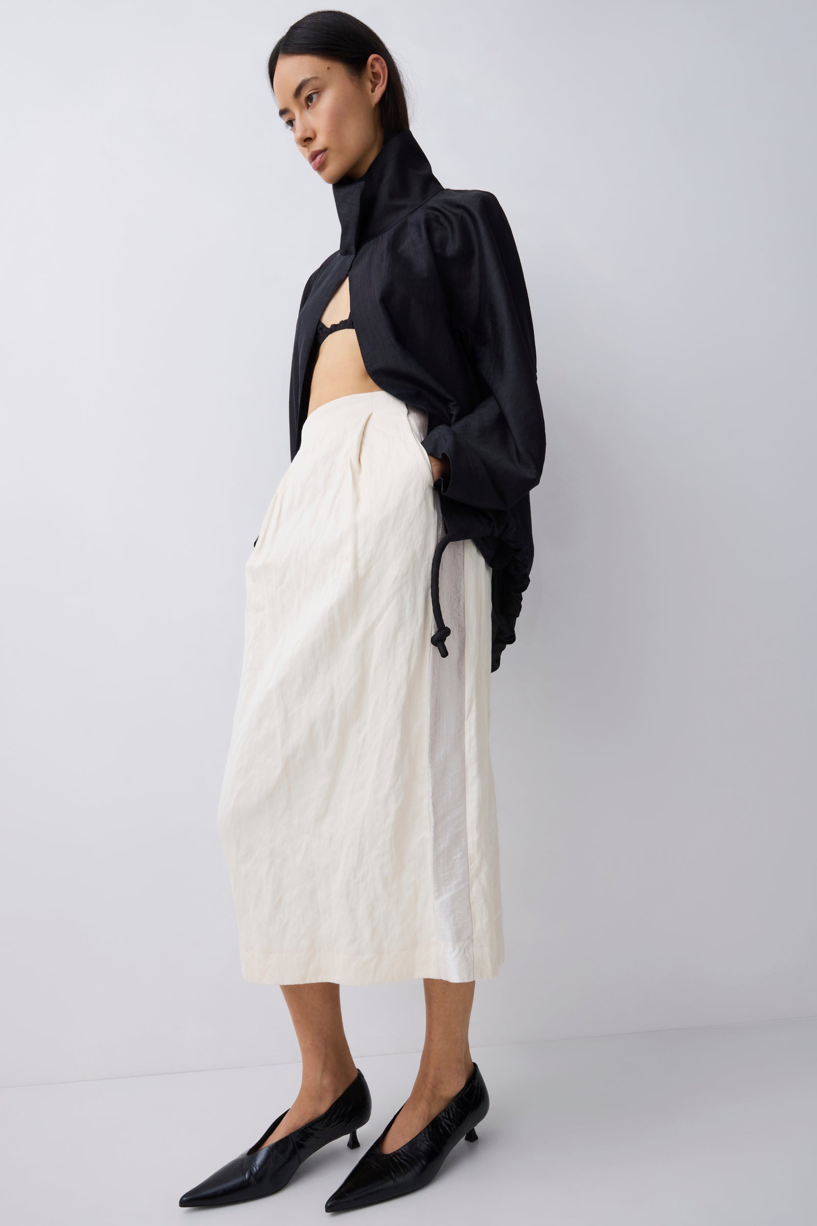 A model wearing a black cropped long-sleeve jacket with a high neck, paired with a cream-colored textured midi skirt and black pointed-toe kitten heels, set against a plain light gray studio background.