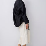 A model is shown from a side-back angle wearing a black oversized long-sleeve top, cream-colored wide-leg cropped trousers, and black pointed-toe kitten heels against a plain light gray studio background.