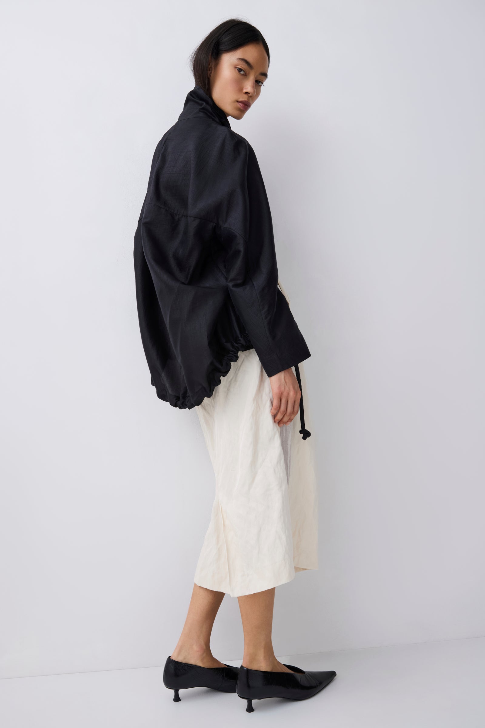 A model is shown from a side-back angle wearing a black oversized long-sleeve top, cream-colored wide-leg cropped trousers, and black pointed-toe kitten heels against a plain light gray studio background.