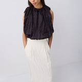 A woman modeling a sleeveless black top with dramatic gathered ruffles and a high neckline, paired with a long cream skirt and black flats, against a plain light gray studio background.