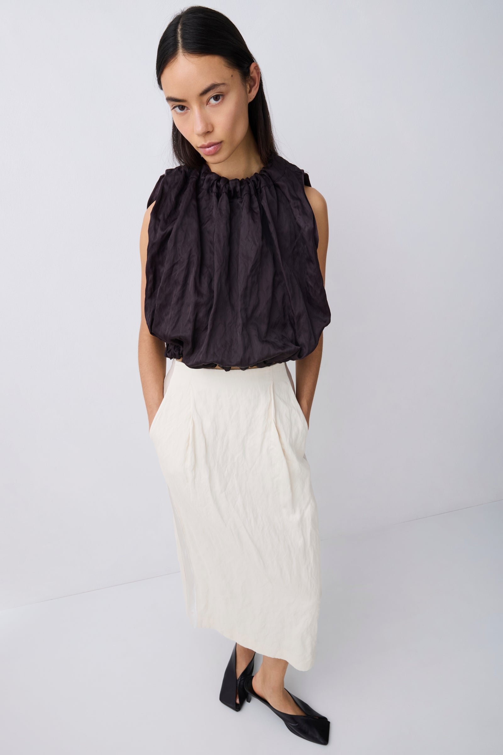 A woman modeling a sleeveless black top with dramatic gathered ruffles and a high neckline, paired with a long cream skirt and black flats, against a plain light gray studio background.