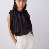 A model wearing a sleeveless black top with a gathered, voluminous ruffled neckline and a white skirt, photographed against a light gray studio background.