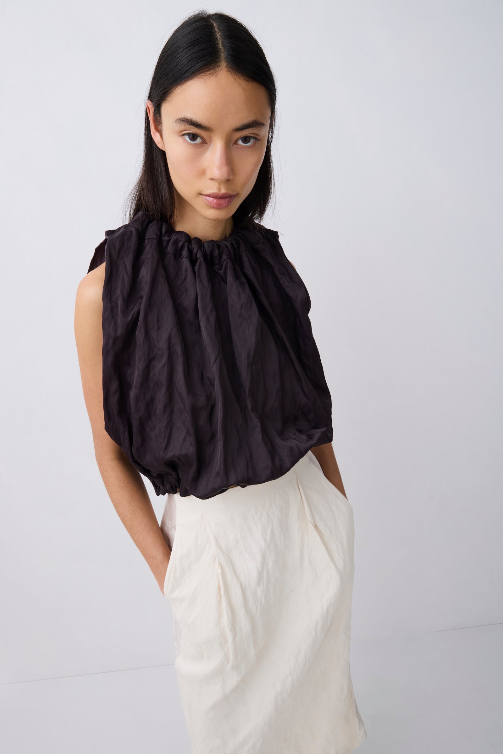 A model wearing a sleeveless black top with a gathered, voluminous ruffled neckline and a white skirt, photographed against a light gray studio background.