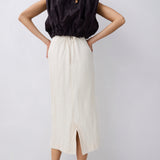A woman shown from the back wearing a black sleeveless top with a gathered back and a long cream midi skirt with a front slit, paired with black pointed heels, against a light gray studio background.