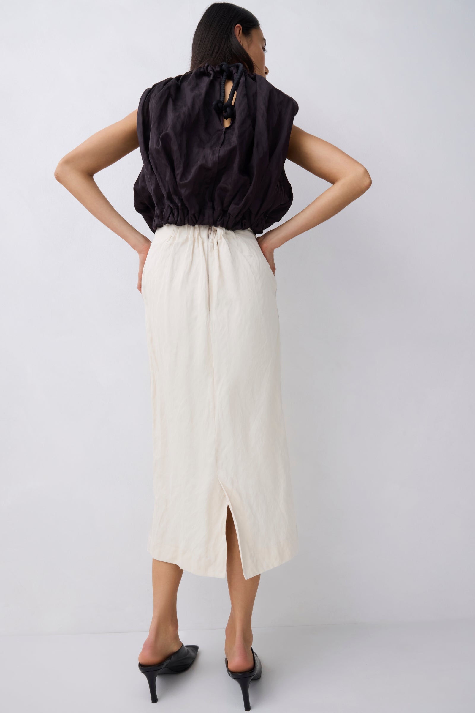 A woman shown from the back wearing a black sleeveless top with a gathered back and a long cream midi skirt with a front slit, paired with black pointed heels, against a light gray studio background.