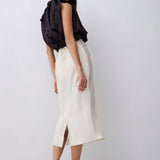 A model is shown from the back wearing a cream-colored midi skirt with a front slit, paired with a black draped top and black mule heels, against a light gray studio background.