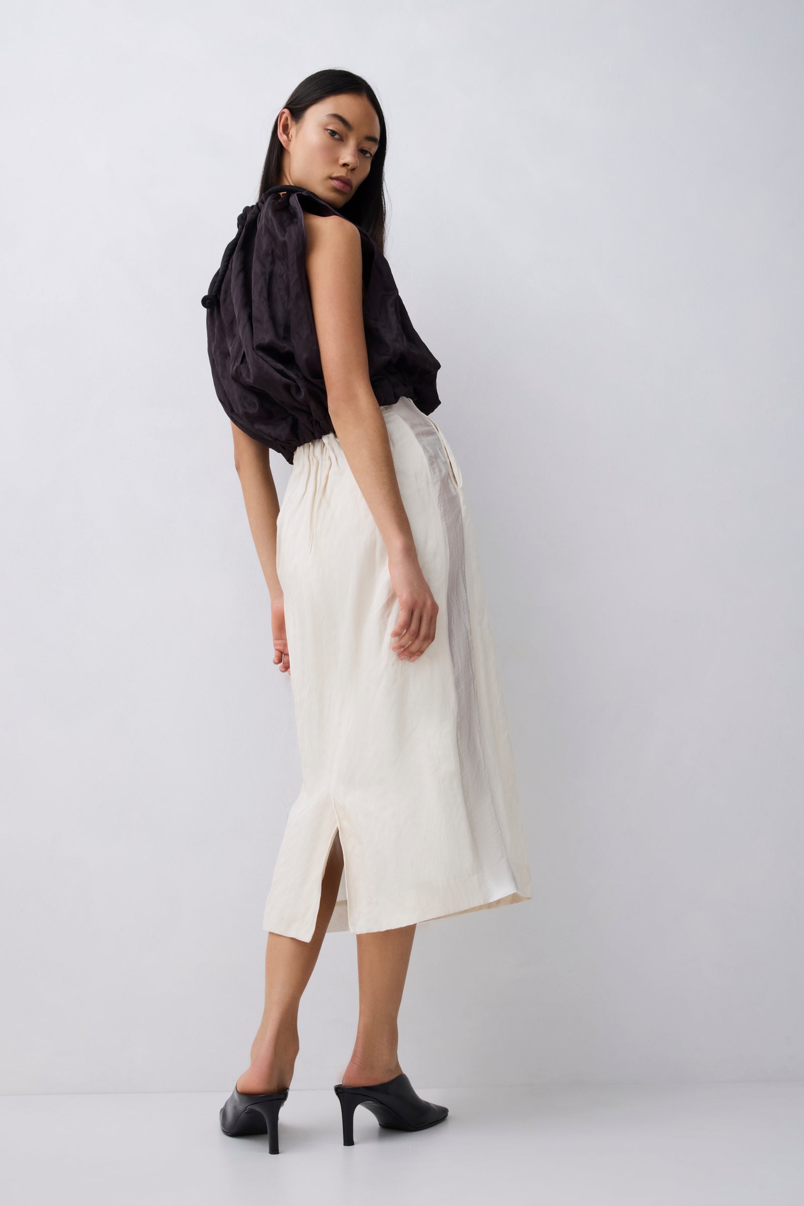A model is shown from the back wearing a cream-colored midi skirt with a front slit, paired with a black draped top and black mule heels, against a light gray studio background.