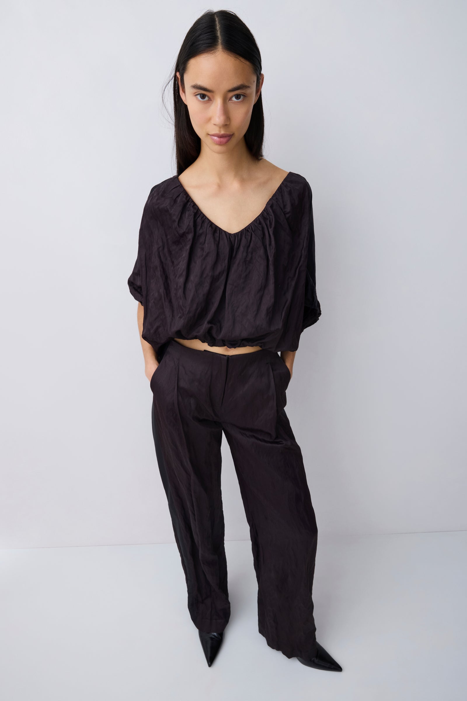 A model wearing a black textured two-piece set consisting of a cropped, wide-sleeved V-neck top and matching wide-leg trousers, photographed against a plain light gray studio background.