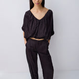 A model wearing a black textured two-piece set consisting of a cropped, wide-sleeved V-neck top and matching wide-leg trousers, photographed against a plain light gray studio background.