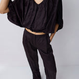 A model wearing a black crinkled two-piece outfit consisting of a cropped, wide-sleeved V-neck top and matching relaxed-fit trousers, paired with black pointed-toe heels against a light gray studio background.