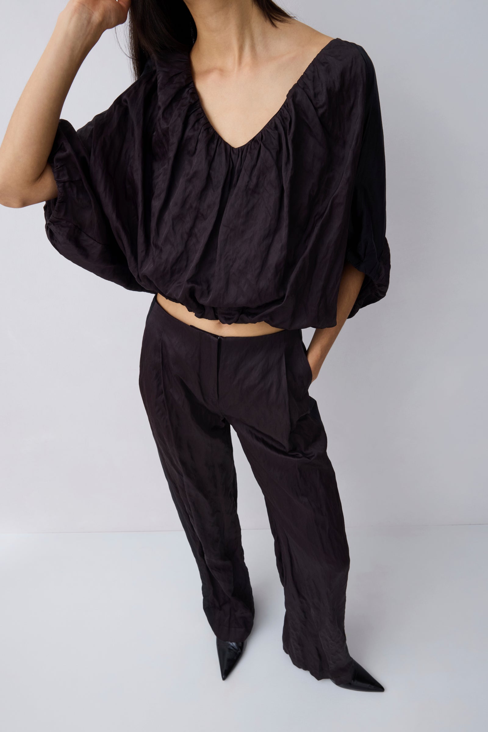 A model wearing a black crinkled two-piece outfit consisting of a cropped, wide-sleeved V-neck top and matching relaxed-fit trousers, paired with black pointed-toe heels against a light gray studio background.