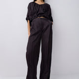 A model stands against a light gray studio background wearing a black cropped top with voluminous short sleeves and matching black wide-leg trousers, paired with black pointed shoes.