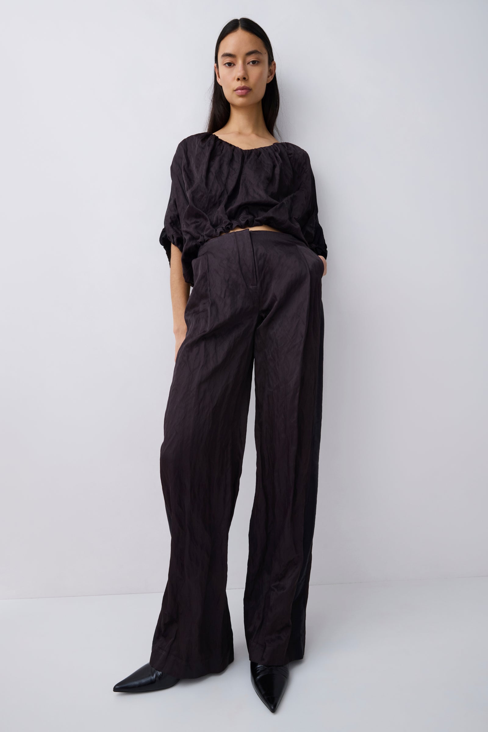 A model stands against a light gray studio background wearing a black cropped top with voluminous short sleeves and matching black wide-leg trousers, paired with black pointed shoes.