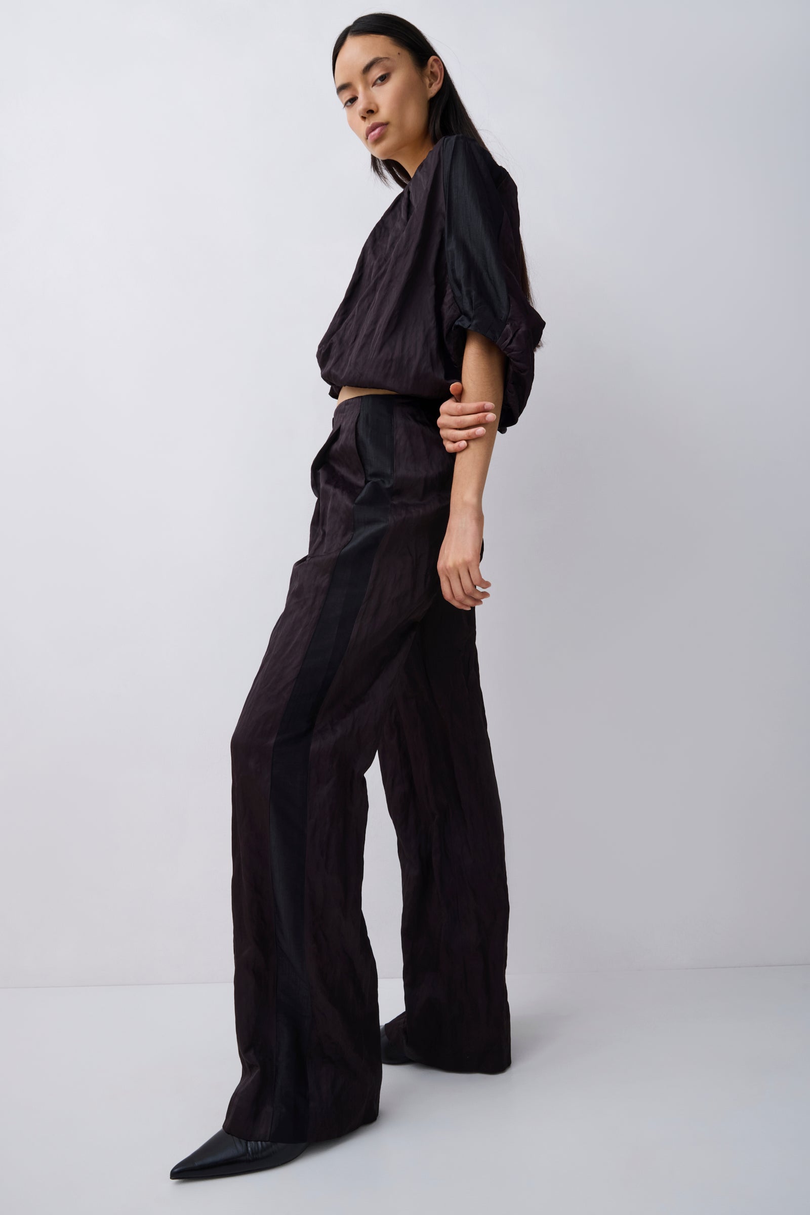 A model wearing a black draped two-piece outfit consisting of a cropped long-sleeve top and matching wide-leg trousers, standing against a light gray studio background.