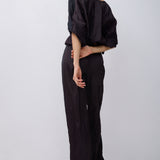 A model is shown from the back wearing a black off-the-shoulder top with voluminous puff sleeves and matching black wide-leg trousers, set against a plain light gray studio background.
