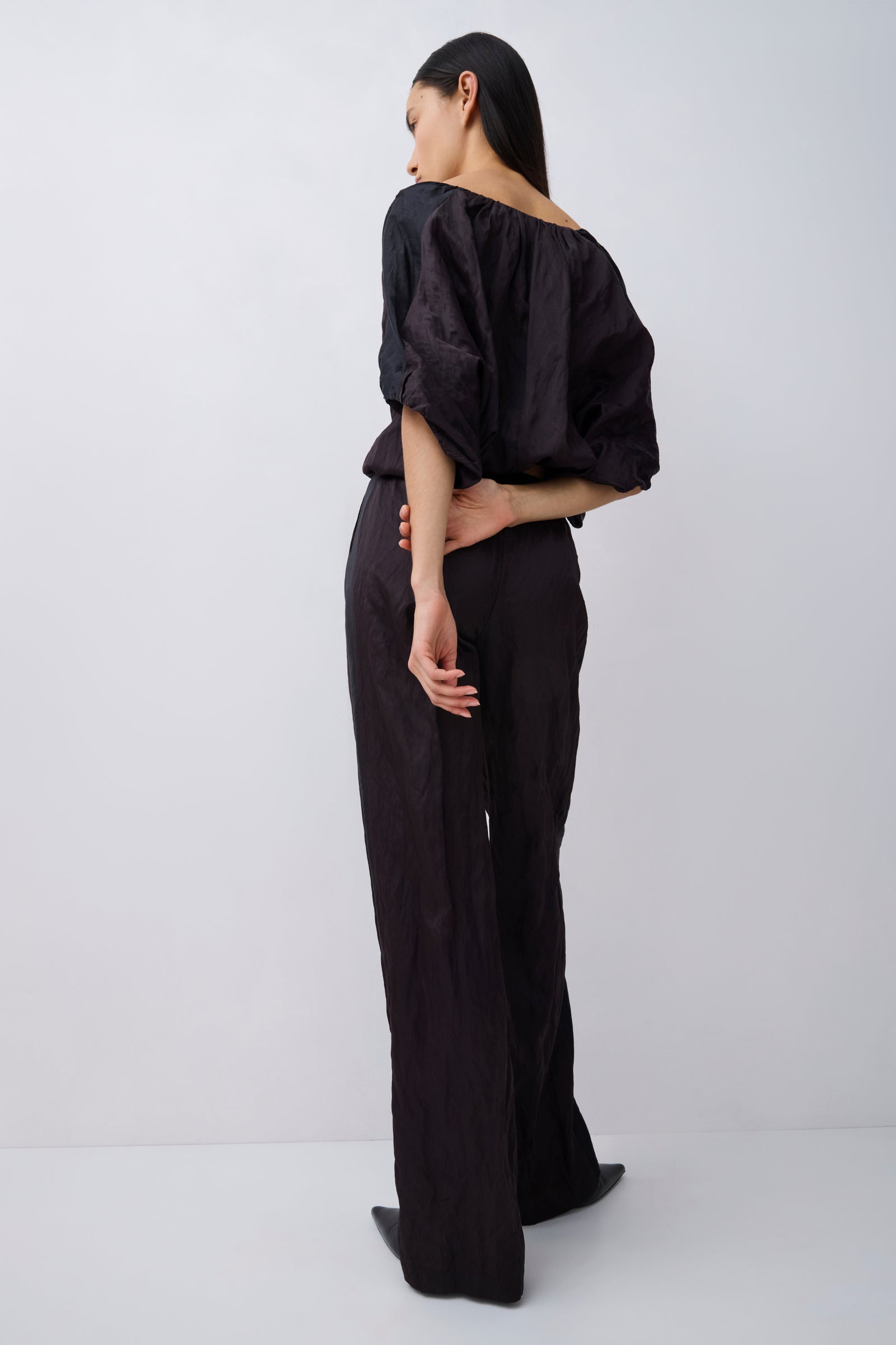 A model is shown from the back wearing a black off-the-shoulder top with voluminous puff sleeves and matching black wide-leg trousers, set against a plain light gray studio background.